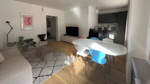 Ett modernt vardagsrum med en soffa, ett matbord med stolar, en TV och en köksdel med vitvaror.