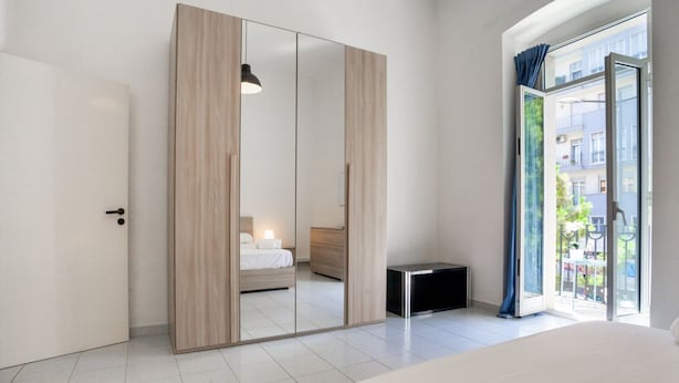 A modern bedroom with a large mirror, a bed, a bedside table, and a sliding door leading to a balcony.