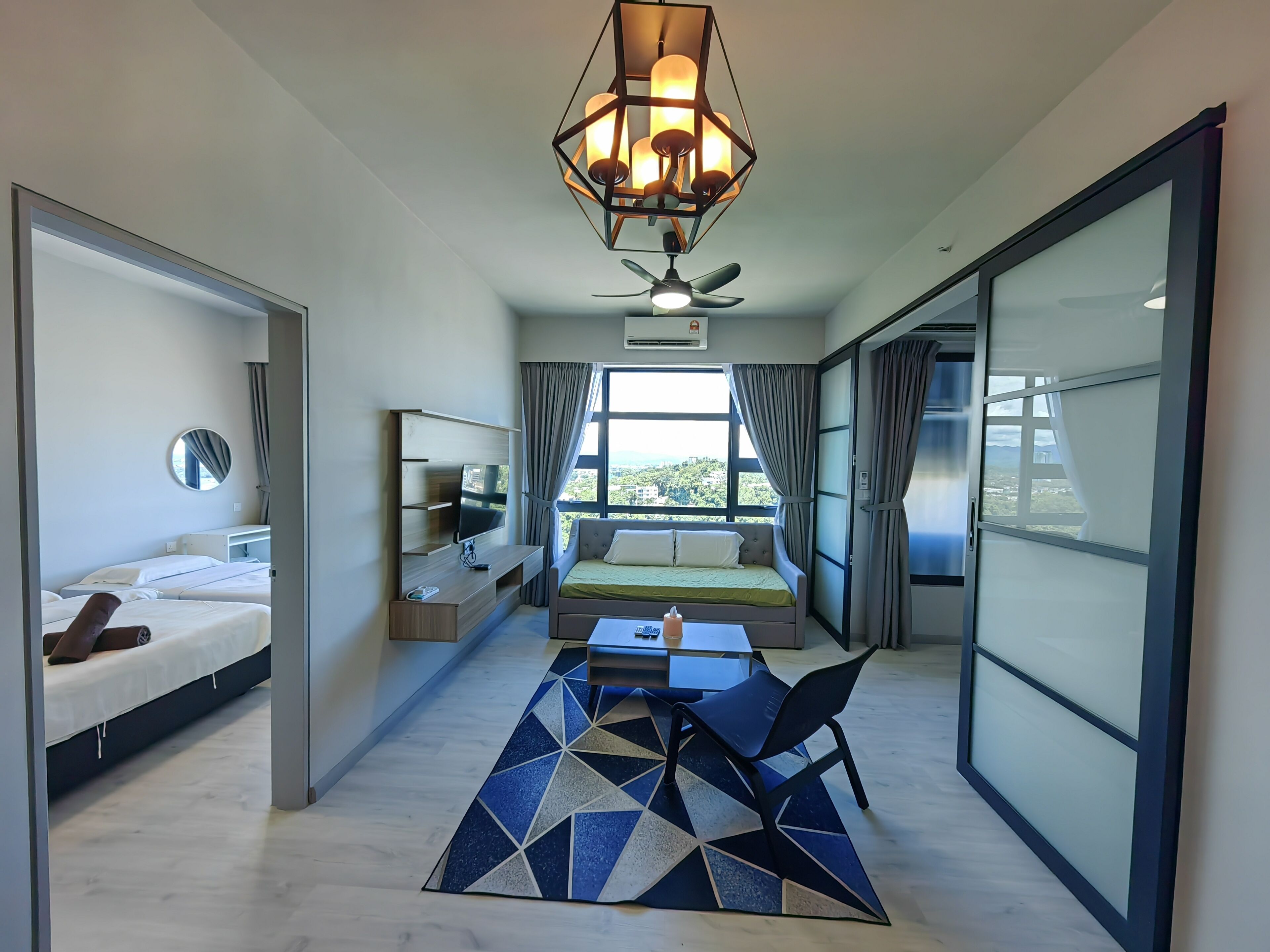 A modern hotel room with a bed, a desk with a computer, a chair, a sofa, a ceiling fan, and a large window offering a city view.
