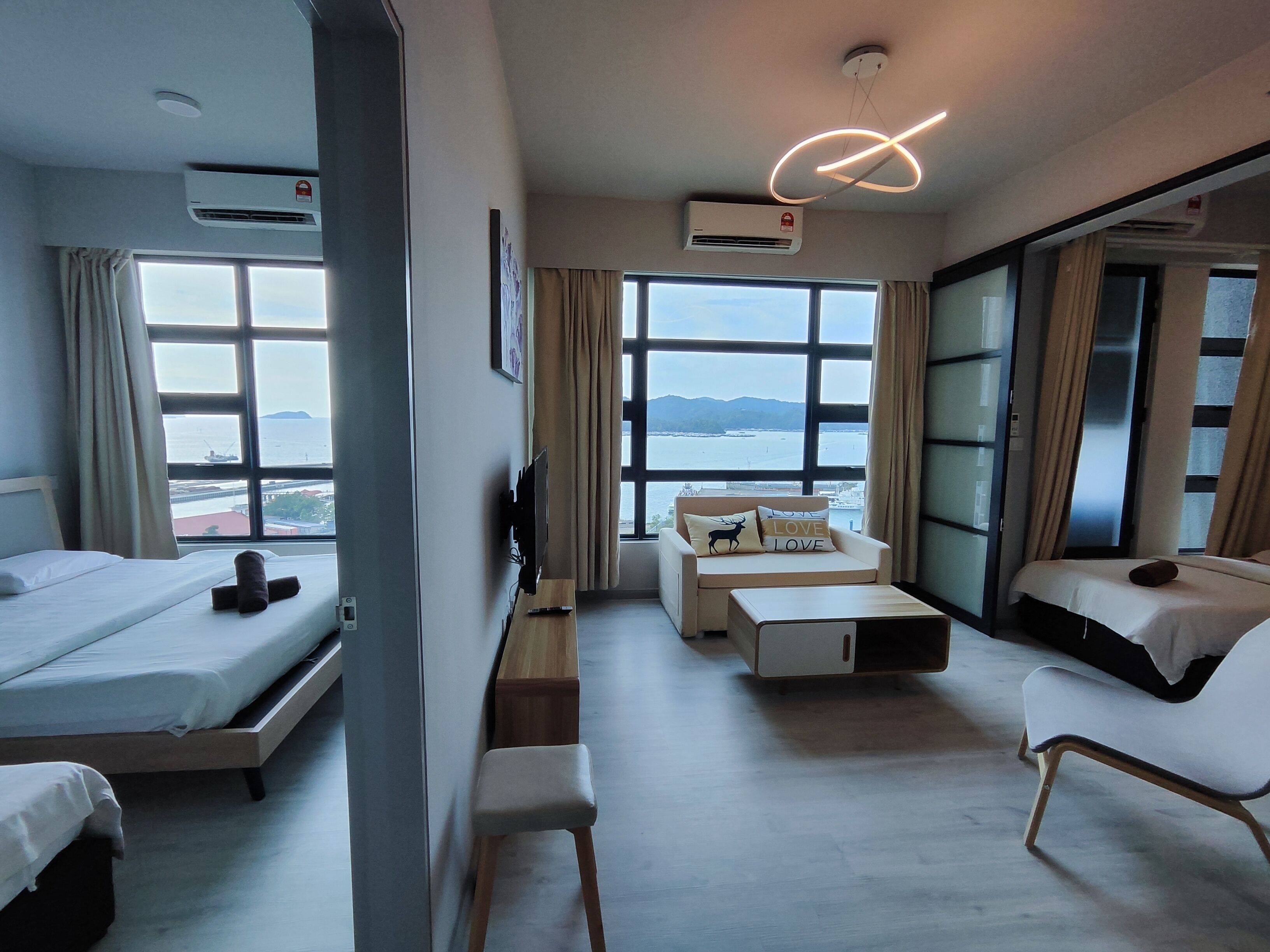 Una habitación de hotel moderna con cama, escritorio, sofá y sillón. Amplias ventanas con vistas al mar y a las montañas.