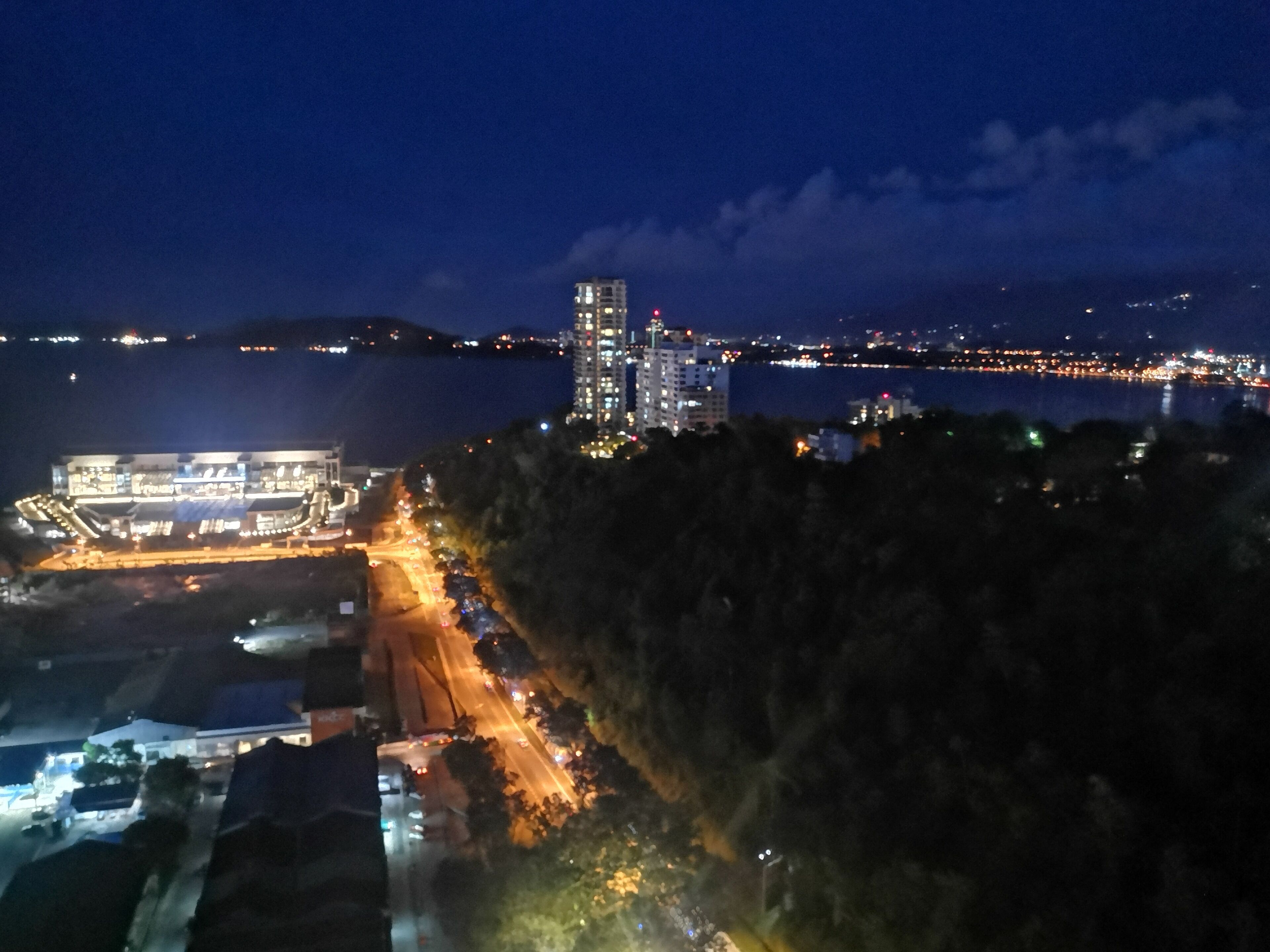 A nighttime view of a coastal cityscape with a prominent high-rise building, a lit-up waterfront, and a road leading to a building complex.