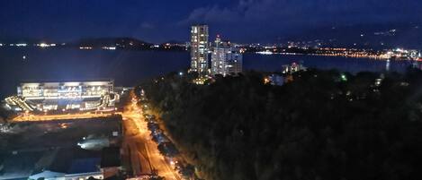 A nighttime view of a coastal cityscape with a prominent high-rise building, a lit-up waterfront, and a road leading to a building complex.