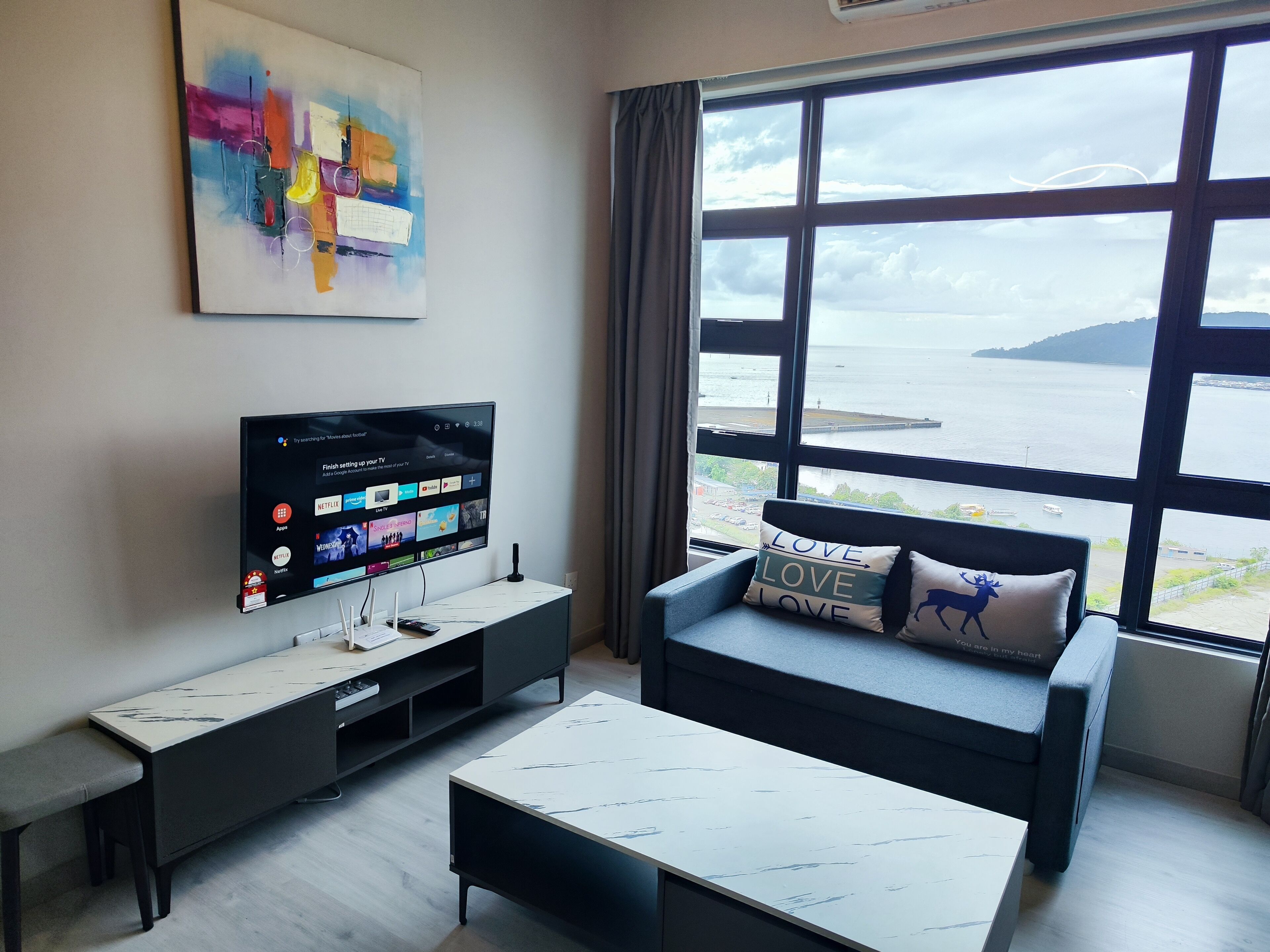 A modern living room with a TV, a sofa with decorative pillows, a coffee table, and a large window offering a view of the sea.
