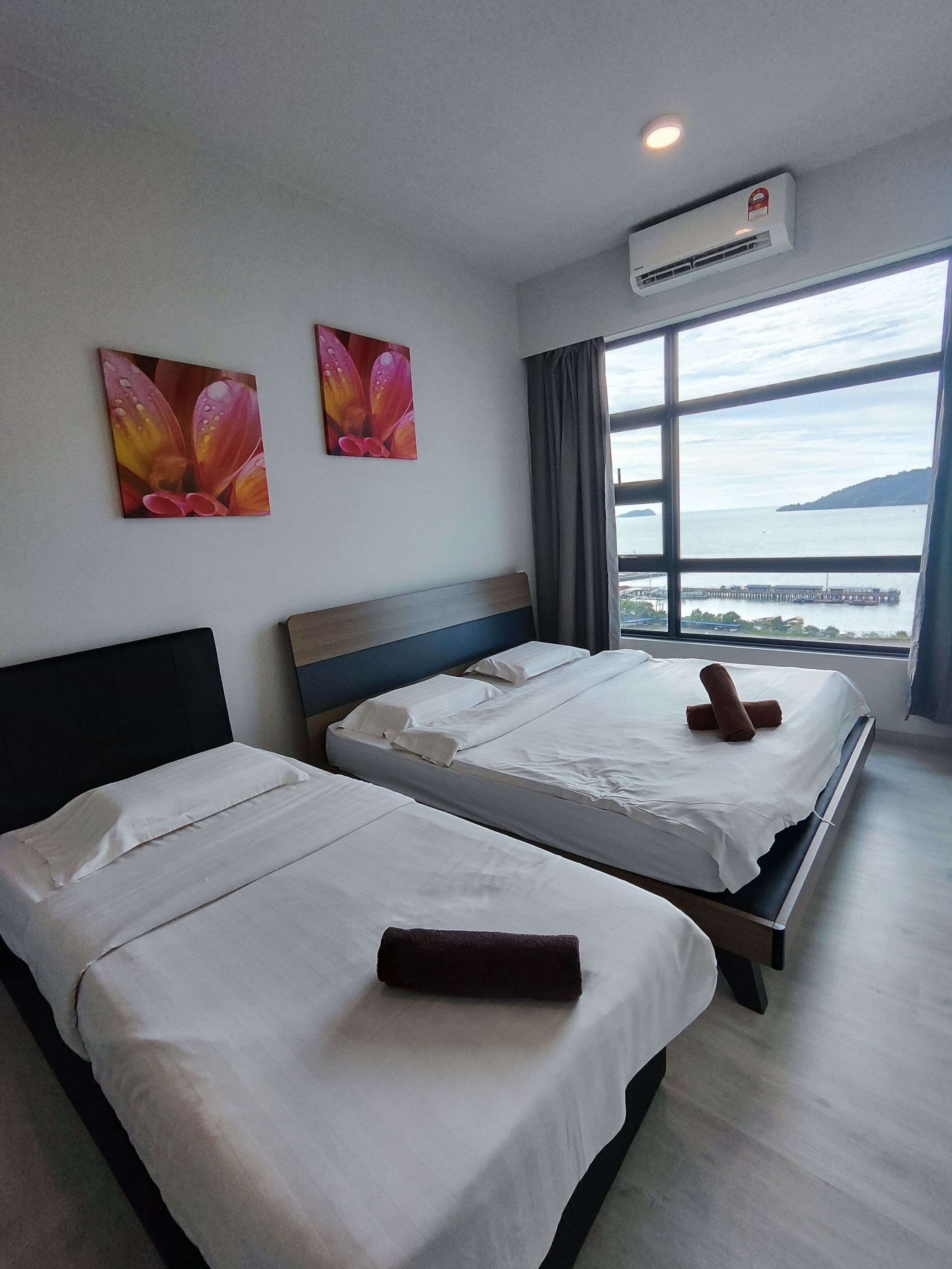 A hotel room with two beds, a large window with a view of a body of water and a pier, and an air conditioning unit on the wall.