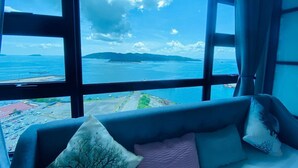 A room with a large window offering a view of a coastal area, a sofa with cushions, and a curtain.