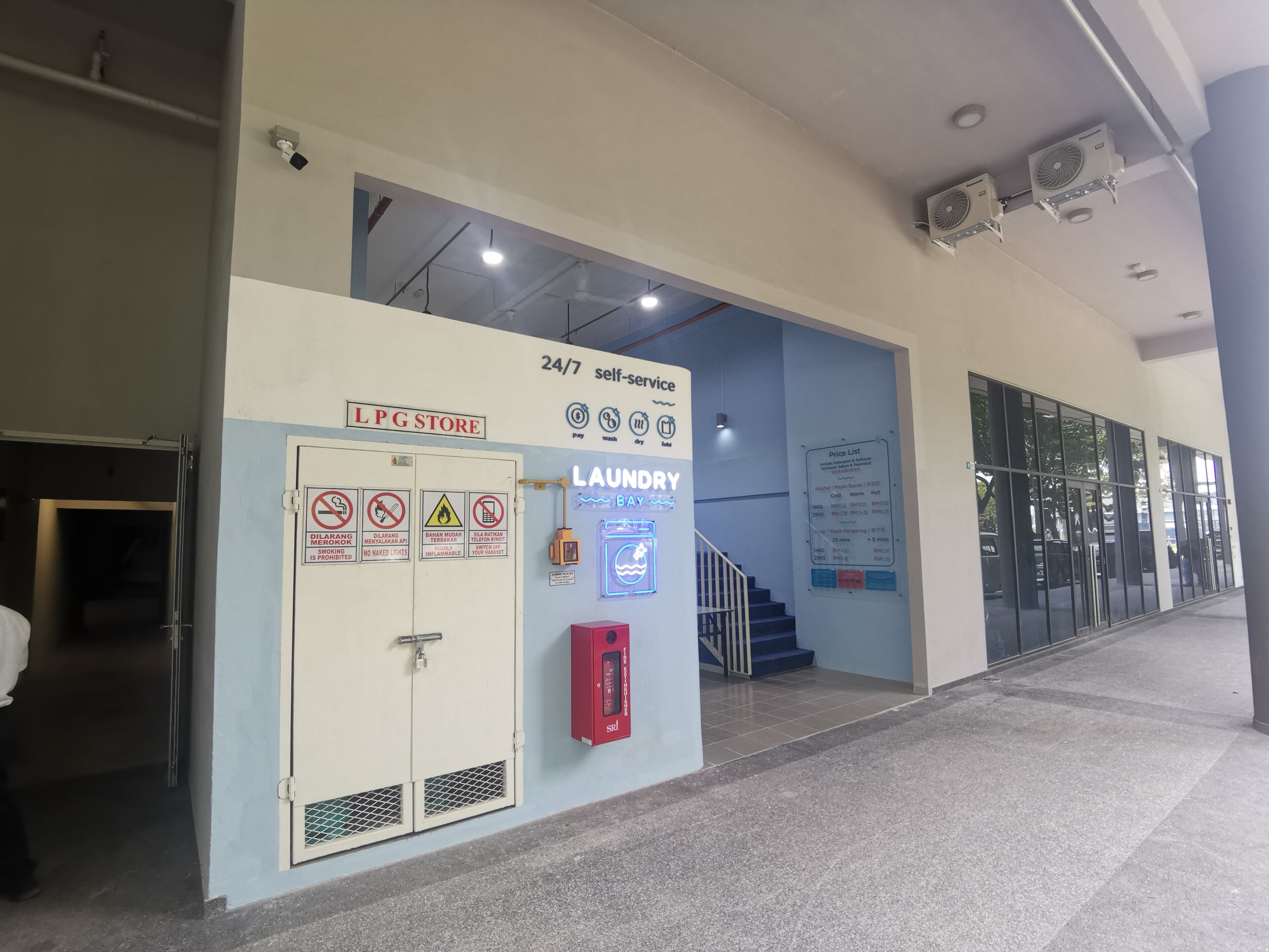 Une laverie en libre-service avec une pancarte indiquant qu’elle est ouverte 24 heures sur 24, 7 jours sur 7, un magasin de vente de bouteilles de gaz et un extincteur.