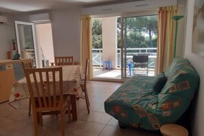 A room with a dining table and chairs, a sofa, a sliding glass door leading to an outdoor area, and a small kitchen area. - Apartment for 4 people, air-conditioned, sleeps 4, 15 m² terrace – beach and city center within walking distance (Mandelieu-la-Napoule)