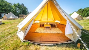 A large, triangular tent with a central bed and a small table, surrounded by grassy area with other tents in the background.