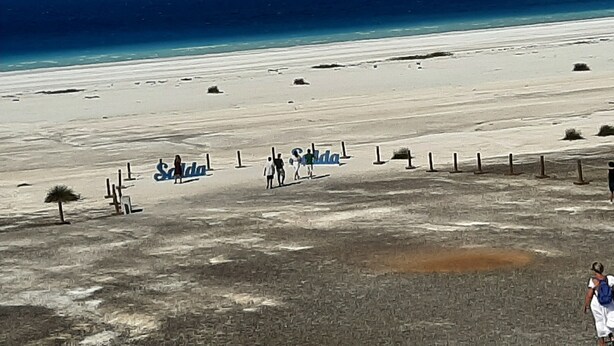Una playa con vista despejada al océano y montañas a lo lejos. Se ven letreros con el nombre 'Saldana'.
