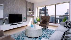 A modern living room with a flat-screen TV, a round coffee table, a grey sofa, and a large window offering a city view.