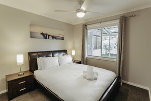 Un lit bien fait, avec du linge de lit blanc, une tête de lit sombre et un ventilateur de plafond.