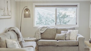 Een woonkamer met een beige bank, een vloerlamp en een groot raam met uitzicht op bomen.