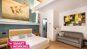 Une chambre moderne avec un grand lit, un canapé gris et des œuvres d’art accrochées aux murs.