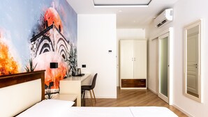 Ein modernes Hotelzimmer mit einem großen Bett, einem Schreibtisch und einem Stuhl. An der Wand hängt ein Gemälde und unter der Decke befindet sich eine Klimaanlage.
