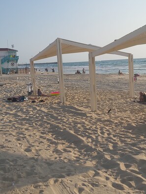Een strand met een uitkijktoren voor de strandwacht, een overdekt zitgedeelte en mensen in de verte.