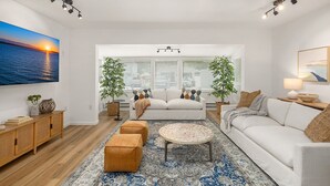 A modern living room with a sofa, ottomans, a coffee table, and a large painting of a sunset. - Coastal Cottage, 4 Large Decks, Close to Beach, Short or Long Stays! (Blaine)