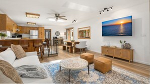 A spacious living area with a dining table, seating, a kitchen, and a large TV displaying a sunset over water. - Coastal Cottage, 4 Large Decks, Close to Beach, Short or Long Stays! (Blaine)