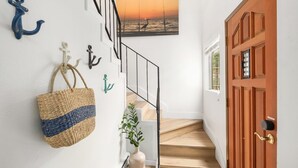 A staircase with a wooden finish, a white wall with various hooks, a woven basket, and a framed picture of a sunset over the ocean. - Coastal Cottage, 4 Large Decks, Close to Beach, Short or Long Stays! (Blaine)