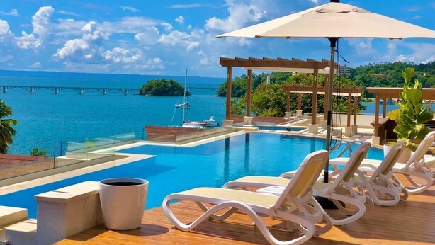 A poolside area with lounge chairs, a pergola, and a view of the ocean and a bridge.