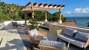 An outdoor seating area with wicker furniture and a pergola, overlooking a body of water and lush greenery.