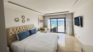 A modern bedroom with a large bed, a sofa, a flat-screen TV, and a view of the sea through a sliding glass door.