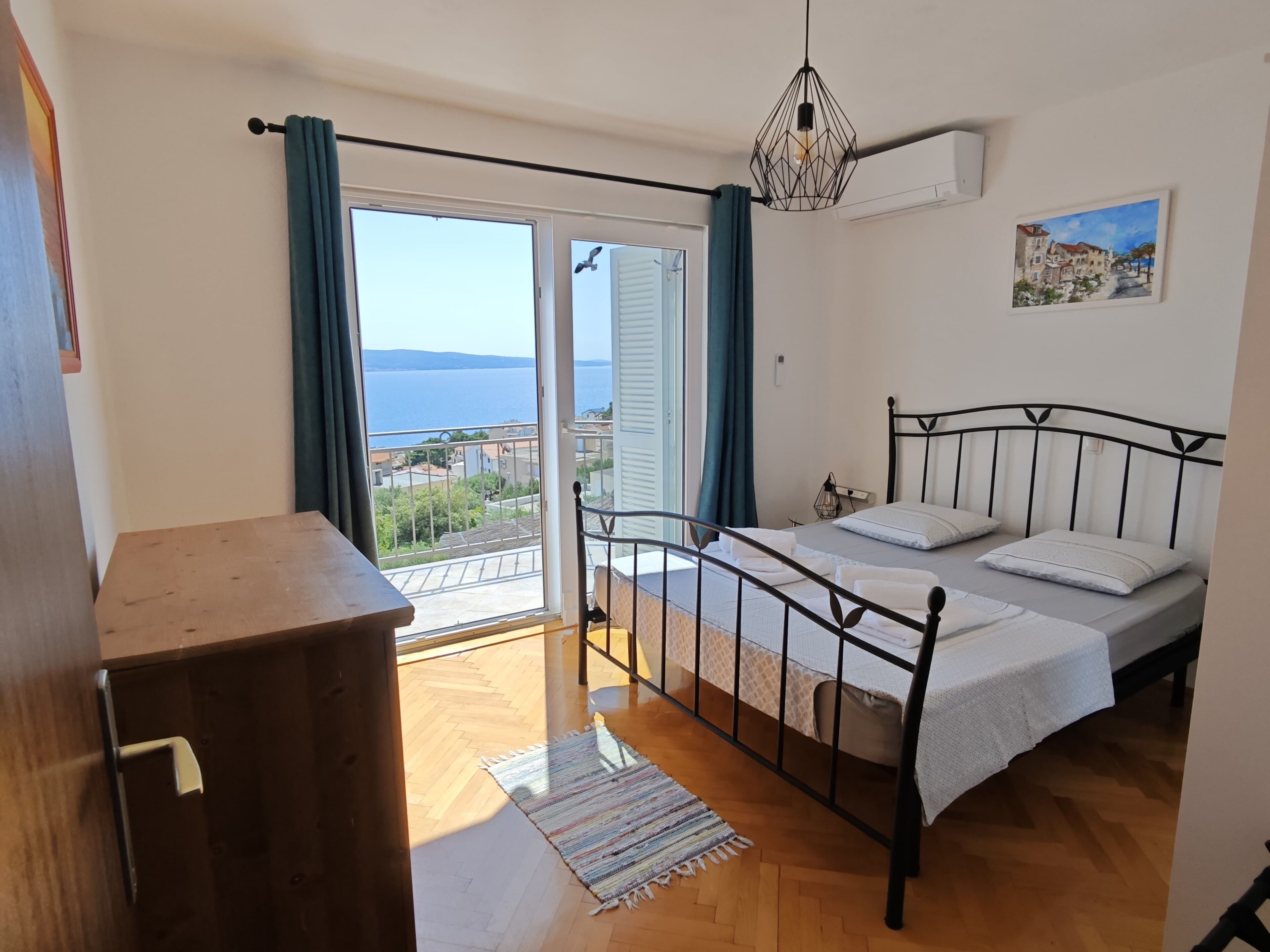 A bedroom with a bed, a wooden dresser, a view of the sea, and a painting on the wall.