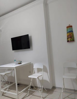 A room with a white desk, two white chairs, a television mounted on the wall, and a colorful artwork on the wall.