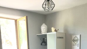Ein Essbereich mit einem Tisch für vier Personen, Holzstühle, einer weißen Tischdecke, einer weißen Kommode mit Ablage und einem Bild im Rahmen an der Wand. Ein Fenster bietet Blick ins Grüne.