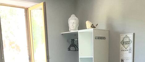 Un comedor con una mesa dispuesta para cuatro, sillas de madera, un mantel blanco, un armario blanco con estante, un cuadro en la pared y una ventana con vistas a follaje.