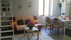 Ein Wohnzimmer mit Sofa, einem kleinen Tisch mit Vase, einem Essbereich mit Stühlen und Tisch sowie einem Bücherregal.
