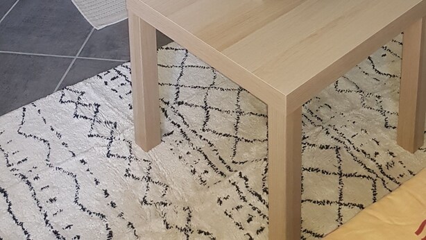 Ein modernes Wohnzimmer mit einer Türkis-farbenen Akzentwand, einem Couchtisch aus Holz mit einer rosa Blumenarragemant, einem kleinen Beistelltisch mit einer Lampe und einer Küchenzeile mit Arbeitsplatte und Schränken.