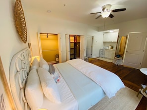 A bedroom with a large bed, a ceiling fan, and a kitchenette in the background.