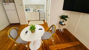 Una cucina compatta con mobili bianchi, un forno a microonde, un fornello e un frigorifero. C'è un'area pranzo con un tavolo bianco e due sedie, una pianta in vaso e un televisore a parete.
