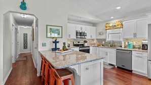 A kitchen with white cabinets, a granite countertop, and a wooden floor. - Happy Honu - Dog-Friendly Beach Retreat, Screened Porch, Walk to Beach (Kure Beach)