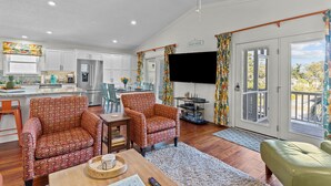A spacious living room with a large TV, two patterned armchairs, a dining area, and a kitchen in the background. - Happy Honu - Dog-Friendly Beach Retreat, Screened Porch, Walk to Beach (Kure Beach)