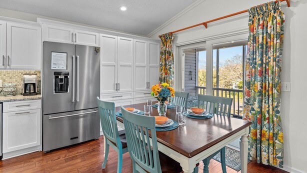 A kitchen with white cabinets, a stainless steel refrigerator, and a dining table set with chairs and a vase of flowers. - Happy Honu - Dog-Friendly Beach Retreat, Screened Porch, Walk to Beach (Kure Beach)