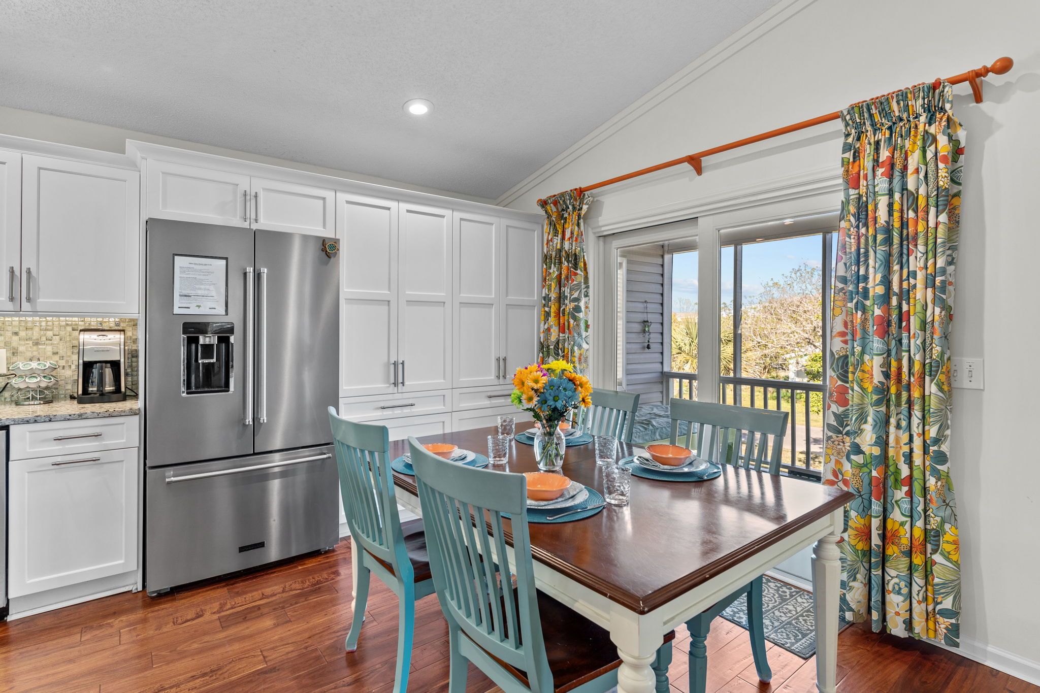 A kitchen with white cabinets, a stainless steel refrigerator, and a dining table set with chairs and a vase of flowers.