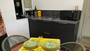 A compact kitchen with a dining area. It includes a small countertop with a microwave, a gas stove, and various kitchen utensils. There are two wicker chairs and a yellow tablecloth-covered table with green plates and cutlery. - Provencal farmhouse, warm and cozy, nestled in the heart of the pool gardens. (Salernes)