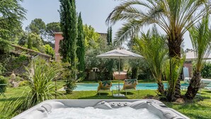 A hot tub with a pool in the background, surrounded by greenery and a chair under a palm tree. - Provencal farmhouse, warm and cozy, nestled in the heart of the pool gardens. (Salernes)