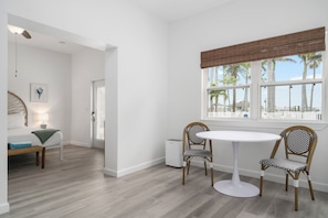 A room with a round white table, two wicker chairs, a bench, a bed, and a window with a view of palm trees. - La Tortuga Historic Downtown Pass A Grille St Pete Beach 6 (St Pete Beach)