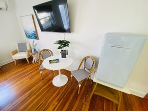 Une pièce de taille modeste avec un plancher en bois, une petite table ronde accompagnée de deux chaises, un téléviseur fixé au mur, un réfrigérateur et un tableau encadré.