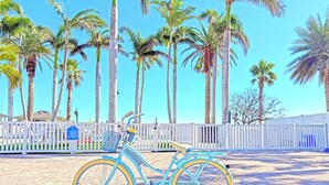 Una bicicleta azul estacionada en una zona pavimentada con una valla blanca y palmeras al fondo.