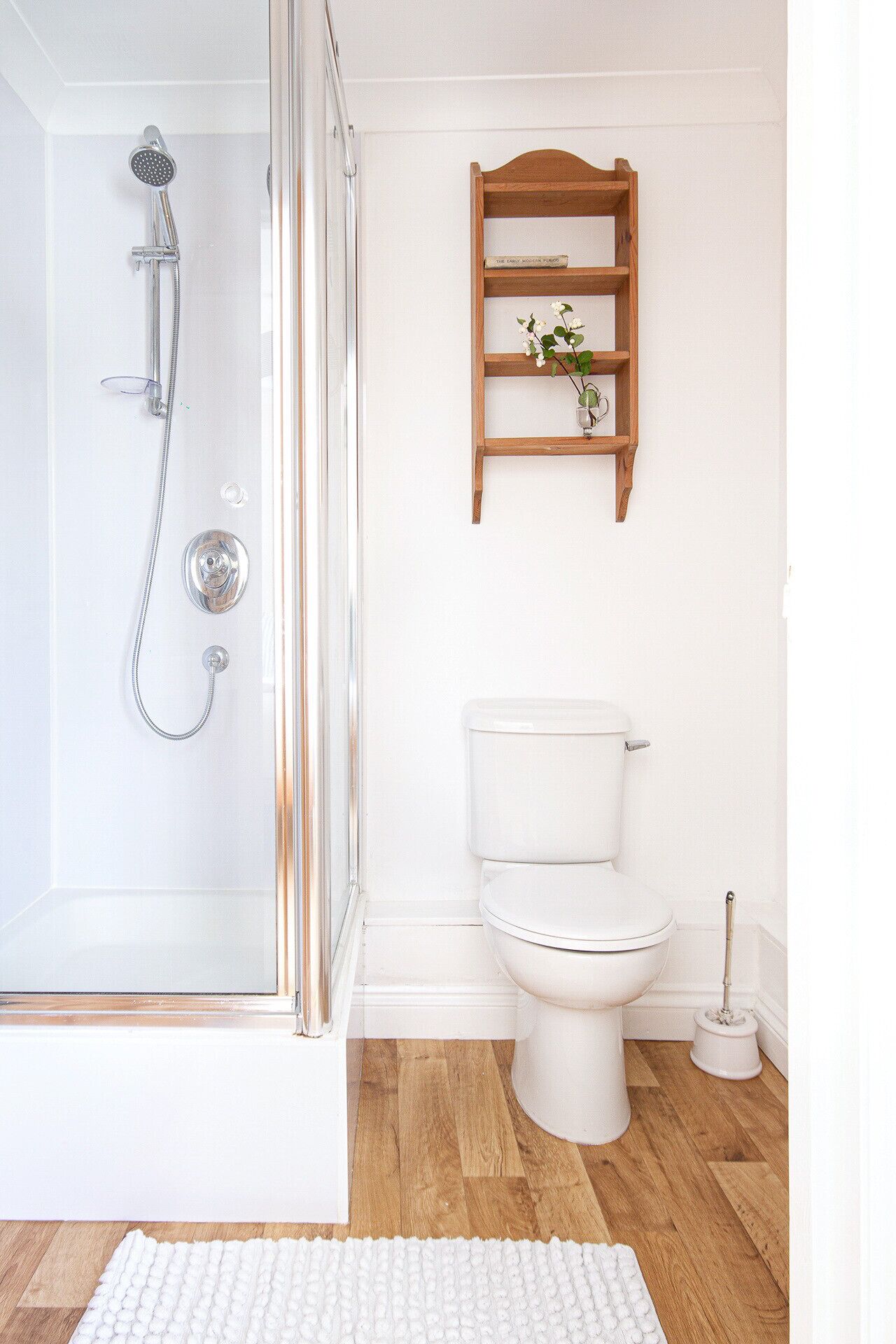 Un baño con ducha de vidrio, inodoro blanco, repisa de madera con planta y alfombra blanca en el suelo.