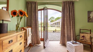 Ein Zimmer mit einem Holzschrank, einer Lampe, einer Vase mit Sonnenblumen, einem Sessel mit Kissen und Blick auf einen Außenbereich durch eine Tür.