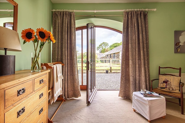 A room with a wooden dresser, a lamp, a vase with sunflowers, a chair with a cushion, and a view of an outdoor area through a doorway. - Gare Barn - Three Bedroom Cottage, Sleeps 6 (Ruan Lanihorne)