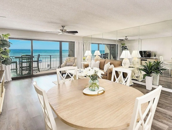 A modern living room with a round wooden dining table, white chairs, a sofa, and a TV. There are potted plants and a view of the ocean through large windows.