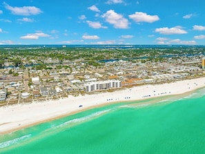 An aerial view of a coastal town with a beachfront hotel, clear turquoise waters, and a dense residential area.