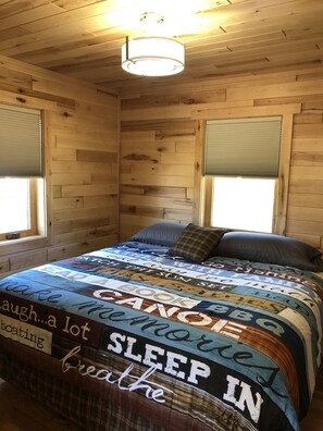 A wooden-paneled room with a bed, a ceiling light, and windows with blinds.
