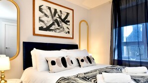 Hotellihuone, jossa on sänky, suuri abstrakti maalaus, peili, ikkuna verhoilla ja yöpöytälamppu.