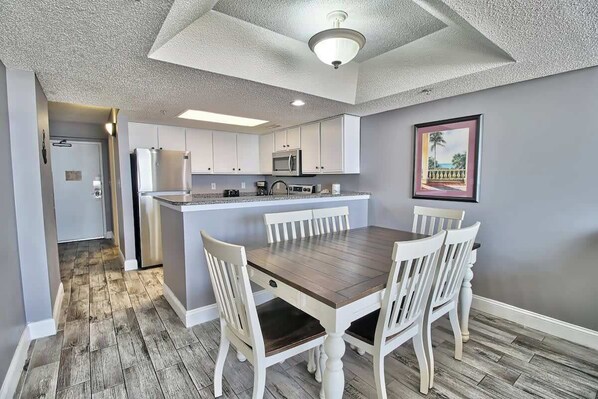 A modern kitchen with white cabinets, a central island, and a dining area with a wooden table and white chairs.